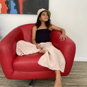 Puede incluir: Una persona joven con un top negro sin mangas y pantalones rosa claro se sienta en un sill&oacute;n rojo. Lleva un sombrero de cubo blanco y verde y mira hacia arriba.