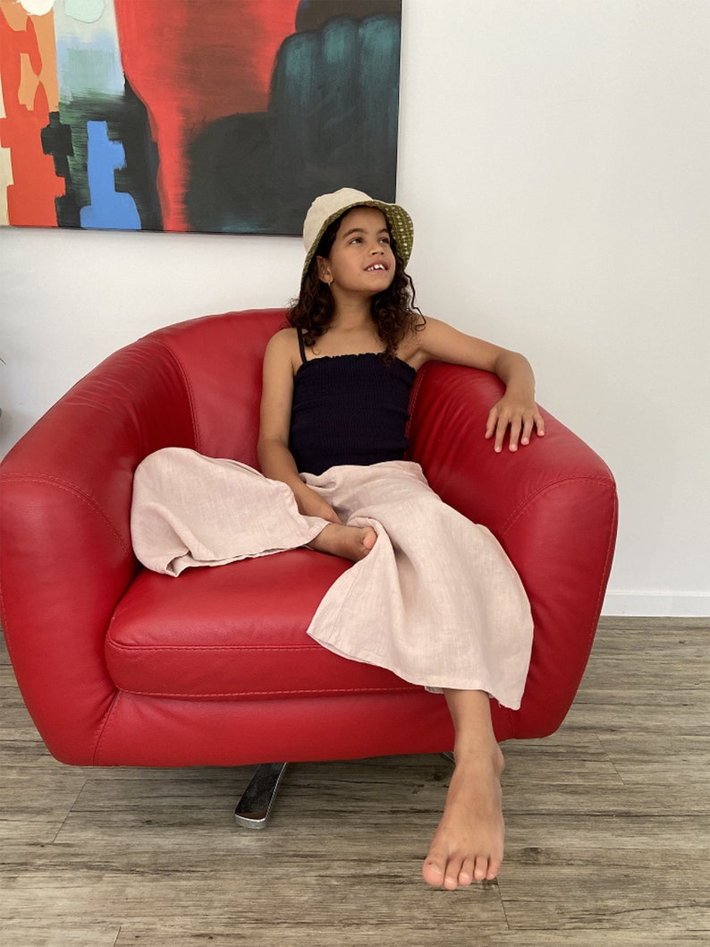 Puede incluir: Una persona joven con un top negro sin mangas y pantalones rosa claro se sienta en un sill&oacute;n rojo. Lleva un sombrero de cubo blanco y verde y mira hacia arriba.