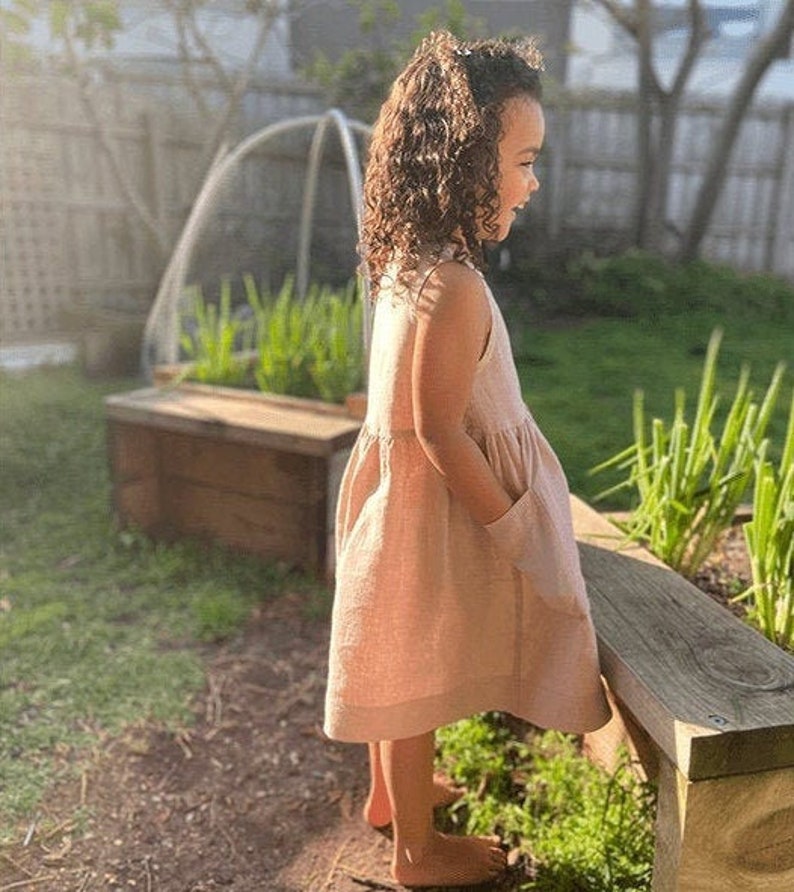 May include: A young girl wearing a light pink sleeveless dress with pockets stands in a garden setting. She is smiling and looking to the right.
