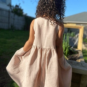May include: A sleeveless, light pink linen dress with a gathered skirt. The dress is flowy and has a casual, relaxed fit.