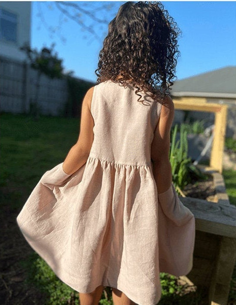 May include: A sleeveless, light pink linen dress with a gathered skirt. The dress is flowy and has a casual, relaxed fit.