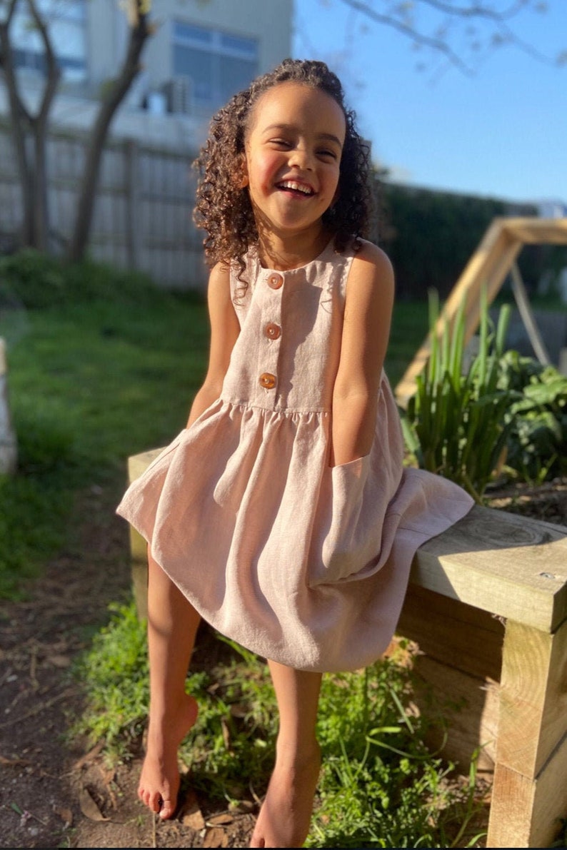 May include: A young girl wearing a sleeveless, light pink linen dress with a button-up front and a pocket on the side. She is sitting on a wooden bench in a garden setting.