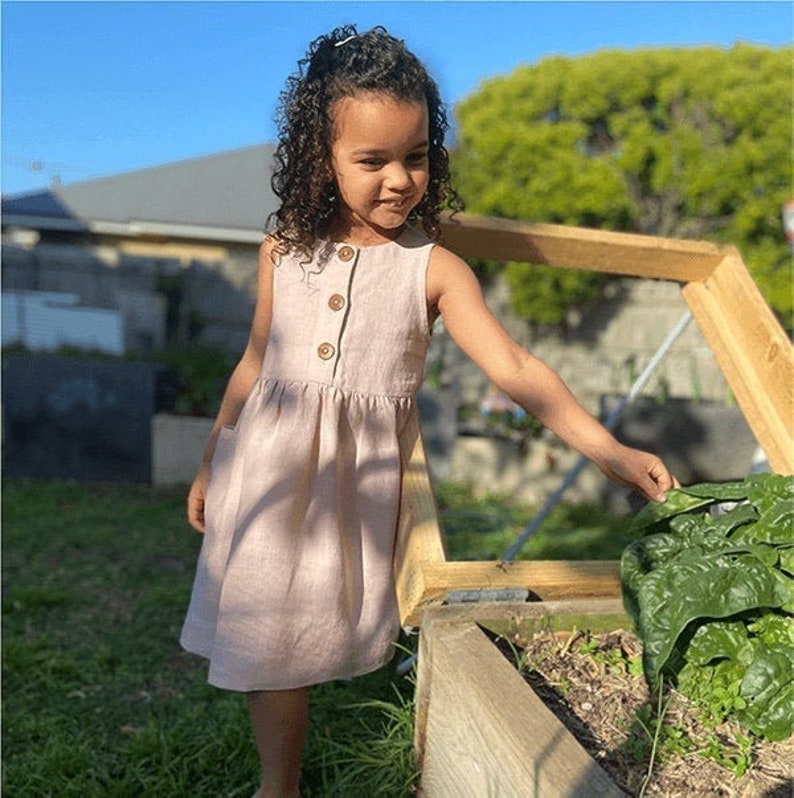 May include: A young girl wearing a light pink linen dress with brown buttons is standing next to a wooden planter box. She is reaching into the planter box to touch a green leafy plant.