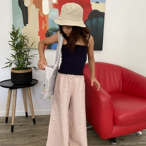 Puede incluir: Una persona joven lleva un top negro sin mangas, pantalones de lino beige y un sombrero de cubo beige. Lleva una bolsa de tela blanca con rayas de arco&iacute;ris.