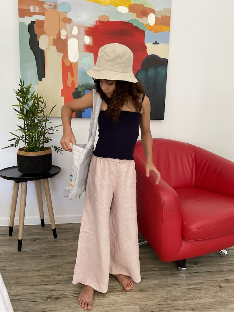 Puede incluir: Una persona joven lleva un top negro sin mangas, pantalones de lino beige y un sombrero de cubo beige. Lleva una bolsa de tela blanca con rayas de arco&iacute;ris.