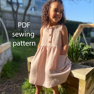 May include: PDF sewing pattern for a sleeveless pink linen dress with a button closure and a pocket. The dress is on a young girl in a garden setting.