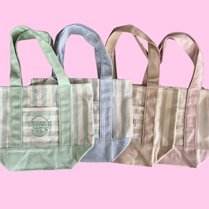 Può includere: Quattro piccole borse tote in tela disposte in fila, ognuna caratterizzata da strisce verticali in colori pastello tra cui verde chiaro, azzurro, beige e rosa. Ogni borsa ha due manici, una tasca frontale e la scritta Trader Joe's ricamata.