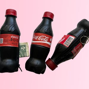 Puede incluir: Tres monederos negros con forma de botella de Coca-Cola con tapas y etiquetas rojas. Un monedero está abierto, revelando una cremallera y un anillo plateado. Un billete de dólar es visible entre dos de los monederos.
