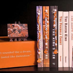 May include: An orange box with a quote and a photo on top, flanked by books and decorative beaded bookmarks. The books have titles like "The Raven King" and "The King's Men". A photo of a football player is on display.