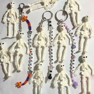 May include: A collection of white plastic skeleton keychains and charms. Some keychains have beaded accents with phrases like "Hocus Pocus", "Stay Spooky", and "Creep It Real". The keychains are arranged on a light-colored surface.