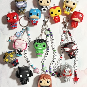 May include: A collection of superhero-themed keychains featuring various characters like Spider-Man, Captain America, and Hulk. Each keychain is a small, colorful figurine attached to a metal clasp and beaded chain, with names spelled out on some chains.