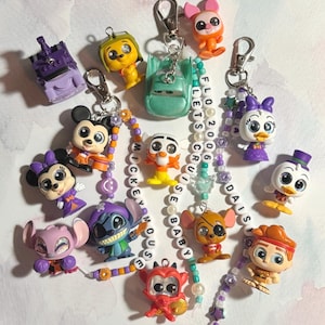 Könnte beinhalten: Eine Sammlung von Schlüsselanhängern mit verschiedenen Cartoon-Figuren, darunter Mickey und Minnie Mouse, Stitch und Donald Duck. Jeder Schlüsselanhänger hat einen silbernen Verschluss und ist an einer Perlenkette mit thematischen Perlen befestigt.