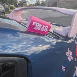 Pode incluir: Um carro azul-escuro com um grande aerofólio traseiro rosa-claro. No aerofólio, há uma placa retangular rosa com o texto branco TOKYO DRIFT. A carroceria é decorada com adesivos de flores de cerejeira em tom rosa-claro.