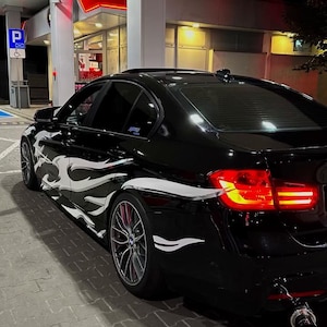 May include: Black car with white flame decals parked under a gas station canopy. The car has tinted windows and custom rims. The word "PEOPLE" is written in white letters at the top of the image.