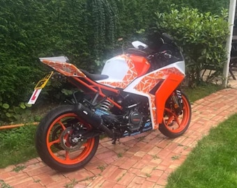 May include: An orange and white KTM RC 390 motorcycle with a black seat and wheels. The motorcycle has a black exhaust and is parked on a brick pathway next to a green lawn and hedge.