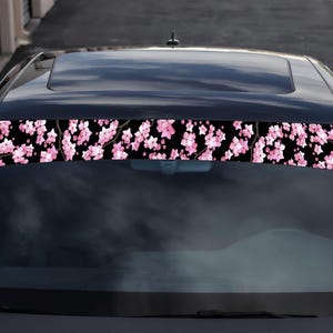 Peut inclure: Un pare-soleil de voiture noir orné d'un motif floral de fleurs de cerisier roses sur fond noir. Les fleurs sont regroupées sur le bord supérieur, créant un accent décoratif pour le pare-brise du véhicule.