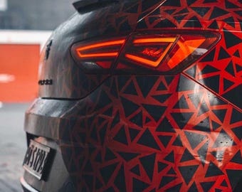May include: Rear view of a car with a red and black geometric pattern. The taillight is illuminated, showing a bright red glow. The car's body has a complex design of black triangles on a red background.