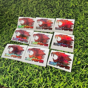 May include: Nine square stickers with a red and orange snarling cat graphic and the text "iBUYPOWER" in white. The stickers have a holographic effect and are arranged on a bed of green grass.