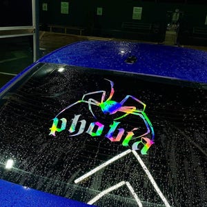 Può includere: Vetro posteriore di un'auto blu con un ragno olografico e la parola "phobia" in gradiente arcobaleno. L'adesivo è applicato sul vetro, con goccioline d'acqua visibili.