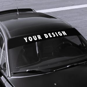 May include: Black car with a windshield banner that reads "YOUR DESIGN" in white block letters. The banner is positioned across the top of the windshield. The car is parked on a gray surface.