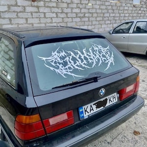 May include: Black BMW station wagon with a white, stylized graphic on the rear window. The car has red taillights and a license plate. The graphic appears to be a decorative sticker or decal.