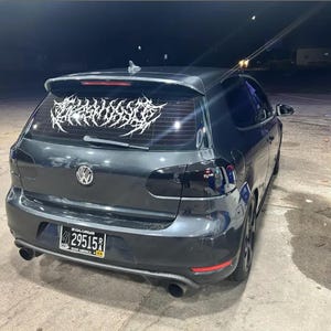 Puede incluir: Hatchback Volkswagen gris oscuro con un gráfico blanco en la luneta trasera. El coche tiene cristales tintados y una matrícula. El coche está aparcado sobre una superficie de hormigón por la noche.
