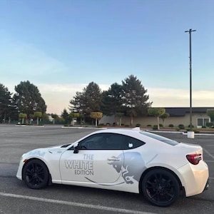 May include: A white two-door coupe car featuring black wheels and a side graphic that includes a stylized bull silhouette and the text THE WHITE EDITION in grey lettering. The vehicle is parked in an outdoor lot under a clear blue sky.