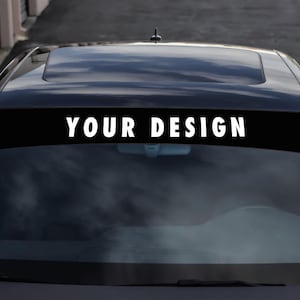 Op de afbeelding: Zwarte voorruitbanner met de tekst "YOUR DESIGN" in het wit, bevestigd aan de bovenkant van de voorruit van een zwarte auto. De banner is een effen zwarte rechthoek en de tekst is vetgedrukt en schreefloos.