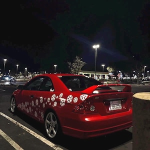 Op de afbeelding: Een rode coupé auto met sneeuwvlokstickers geparkeerd op een parkeerplaats 's nachts. De auto heeft een spoiler en zilveren velgen. De kentekenplaat luidt 9BE2556. De achtergrond toont straatverlichting en andere voertuigen.