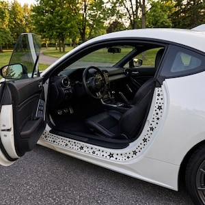 Peut inclure: Voiture de sport blanche avec des autocollants d'étoiles noires sur le cadre de la porte. La portière de la voiture est ouverte, révélant un intérieur noir avec un volant et des sièges. La voiture est garée sur une route avec des arbres en arrière-plan.