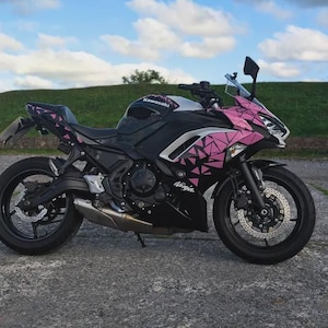 Może przedstawiać: Czarno-różowy motocykl Kawasaki Ninja z geometrycznymi różowymi akcentami. Motocykl ma czarne koła, srebrny zbiornik paliwa, a na boku widnieje napis "Ninja".