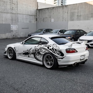 Può includere: Auto sportiva bianca con un design tribale nero sul lato. L'auto ha uno spoiler posteriore e cerchi personalizzati. Diverse altre auto sono parcheggiate sullo sfondo contro un muro di cemento.