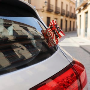 以下が含まれることがあります： 白い車のリアウィンドウに貼られた白、赤、黒のステッカー。ステッカーには太陽光線のような模様と、太い黒のアルファベット大文字でMADE IN JAPANと書かれています。デザインは斜めに配置され、ガラス面で部分的に折れ曲がっています。