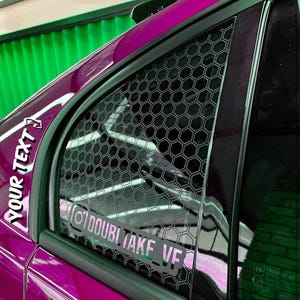 May include: A close-up of a magenta car window with a black honeycomb pattern overlay. The window features a sticker with the text "YOUR TEXT" and another sticker with the Instagram handle "@DOUBLETAKE_VF". The car's paint is glossy.