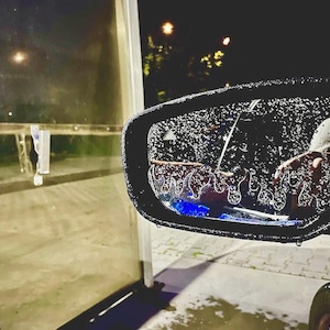 Puede incluir: Primer plano del espejo retrovisor lateral de un coche cubierto de gotas de agua. El espejo refleja una escena oscura con farolas y un vistazo al interior del coche. El espejo tiene un diseño de llama.