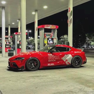 Op de afbeelding: Een felrode sportwagen geparkeerd bij een tankstation in de nacht. De zijkant van het voertuig is voorzien van witte grafische stickers met de tekst THE SUPRA EDITION MKV SERIES en een gestileerde stier-illustratie. Zwarte velgen en rode remklauwen.