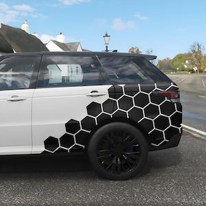 Op de afbeelding: Witte SUV met een zwart honingraatpatroon op de zijkant. Het voertuig heeft zwarte wielen en getinte ramen. De achtergrond toont een straat, huizen en een blauwe lucht.
