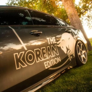 Op de afbeelding: Een donkergrijze sedan met een witte afbeelding van een stier en de tekst "THE KOREAN EDITION" op het zijportier. Het voertuig heeft getinte ruiten, zilverkleurige meerspaaks wielen en een verlaagde stand, gezien vanuit een laag standpunt.