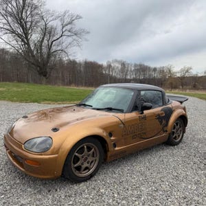 May include: A metallic bronze two-door convertible sports car with a black roof and spoiler. The side panel features a black bull graphic and the text THE AMBER EDITION. The vehicle has dark-colored multi-spoke wheels and a low-profile body design.