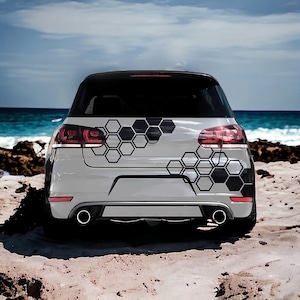May include: Rear view of a white car with a black honeycomb pattern decal. The car has red taillights and dual exhaust pipes. The background shows a beach with sand and ocean.