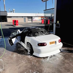 Puede incluir: Un coche descapotable blanco con techo negro y puertas abiertas se está lavando. El coche tiene una calcomanía de llama en la ventana trasera y una matrícula amarilla. El coche está aparcado sobre una superficie de hormigón.