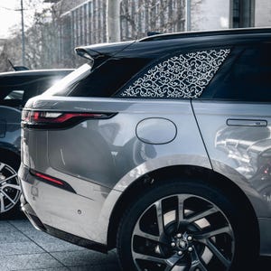 Könnte beinhalten: Ein silberner SUV mit schwarzem Dach und dekorativem weißem Rankenmuster auf der Heckscheibe. Das Fahrzeug hat schwarze Räder und einen schwarzen Spoiler. Das Bild ist im Freien aufgenommen, mit einem Blick auf ein anderes Auto im Hintergrund.