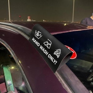Peut inclure: Une étiquette noire pour l'entretien de lavage de voiture avec du texte et des symboles blancs indiquant "LAVAGE À LA MAIN SEULEMENT!". L'étiquette montre un symbole de lavage à la main, un symbole de ne pas sécher en machine et un symbole de ne pas nettoyer à sec.