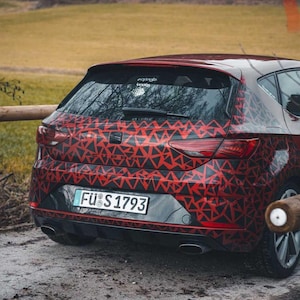 Può includere: Vista posteriore di un'auto rossa con un motivo geometrico nero e rosso. La targa mostra "FÜ S 1793". L'auto è parcheggiata su uno sterrato con una staccionata in legno sullo sfondo.