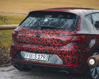 May include: A red hatchback car with a black and red geometric pattern. The car has a license plate that reads "FÜ S 1793". The car is parked on a dirt road with a wooden fence in the background.