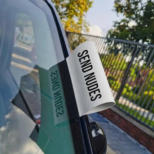 May include: A white sticker with the words "SEND NUDES" in black text is affixed to a car window. The sticker is partially peeled back, revealing the text and its reflection. The image is taken outdoors.