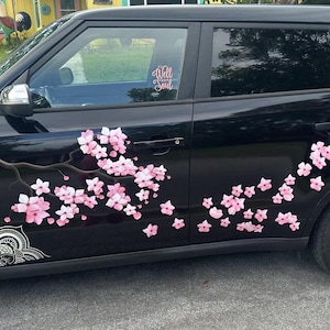 May include: A black car decorated with a large decal of pink cherry blossom flowers on a dark brown branch extending across the side doors. A white mandala-style pattern is at the bottom left, and the text It Is Well With My Soul is on the window.