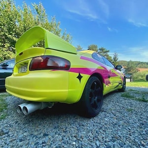 Peut inclure: Une voiture de sport jaune vif avec un grand aileron arrière et des roues noires. La voiture présente des rayures roses et jaunes et des décalcomanies d'étoiles noires. Les tuyaux d'échappement sont visibles. La voiture est garée sur une allée de gravier sous un ciel bleu.