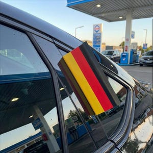 Könnte beinhalten: Ein schwarzes Auto mit einem Aufkleber der deutschen Flagge auf der Scheibe. Die Flagge zeigt schwarze, rote und gelbe vertikale Streifen. Im Hintergrund ist eine Tankstelle mit blau-weißen Schildern zu sehen.