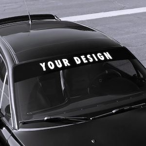 Op de afbeelding: Zwarte auto met een voorruitbanner met de witte tekst "YOUR DESIGN". De banner bevindt zich bovenaan de voorruit. De auto staat op een grijs oppervlak.